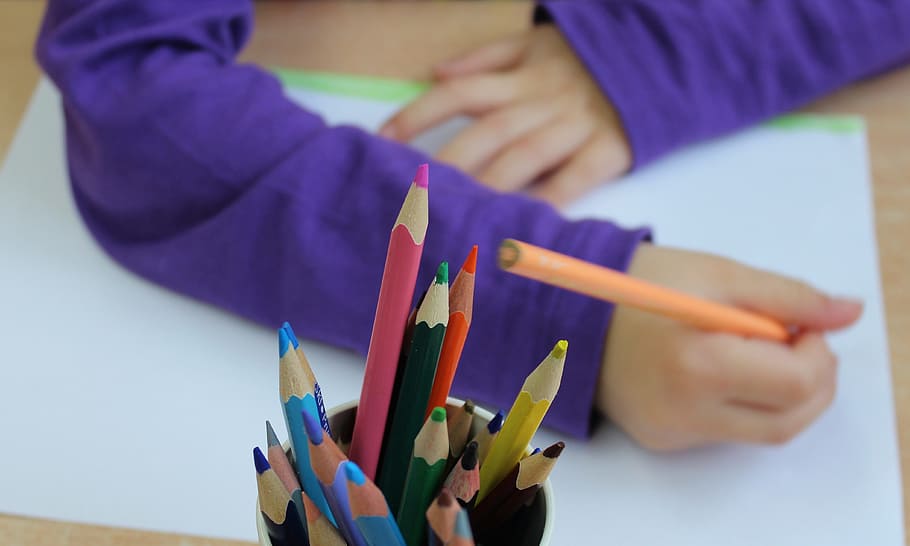 Child's hand with pencil poised over drawing pad.  Jar of colored pencils in foreground.